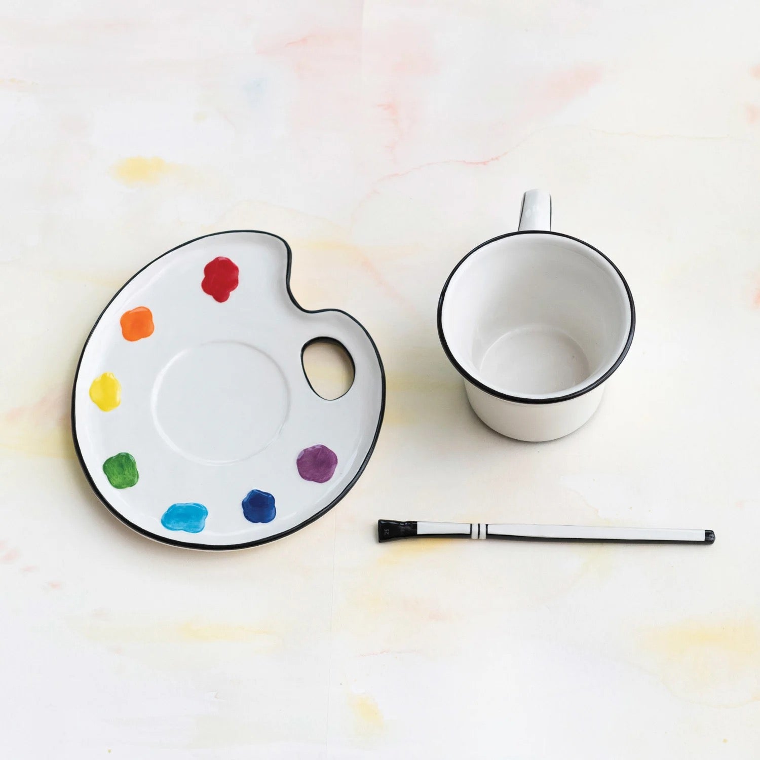 White mug with black rim on a white surface next to a paint palette with colorful dots and a brush.