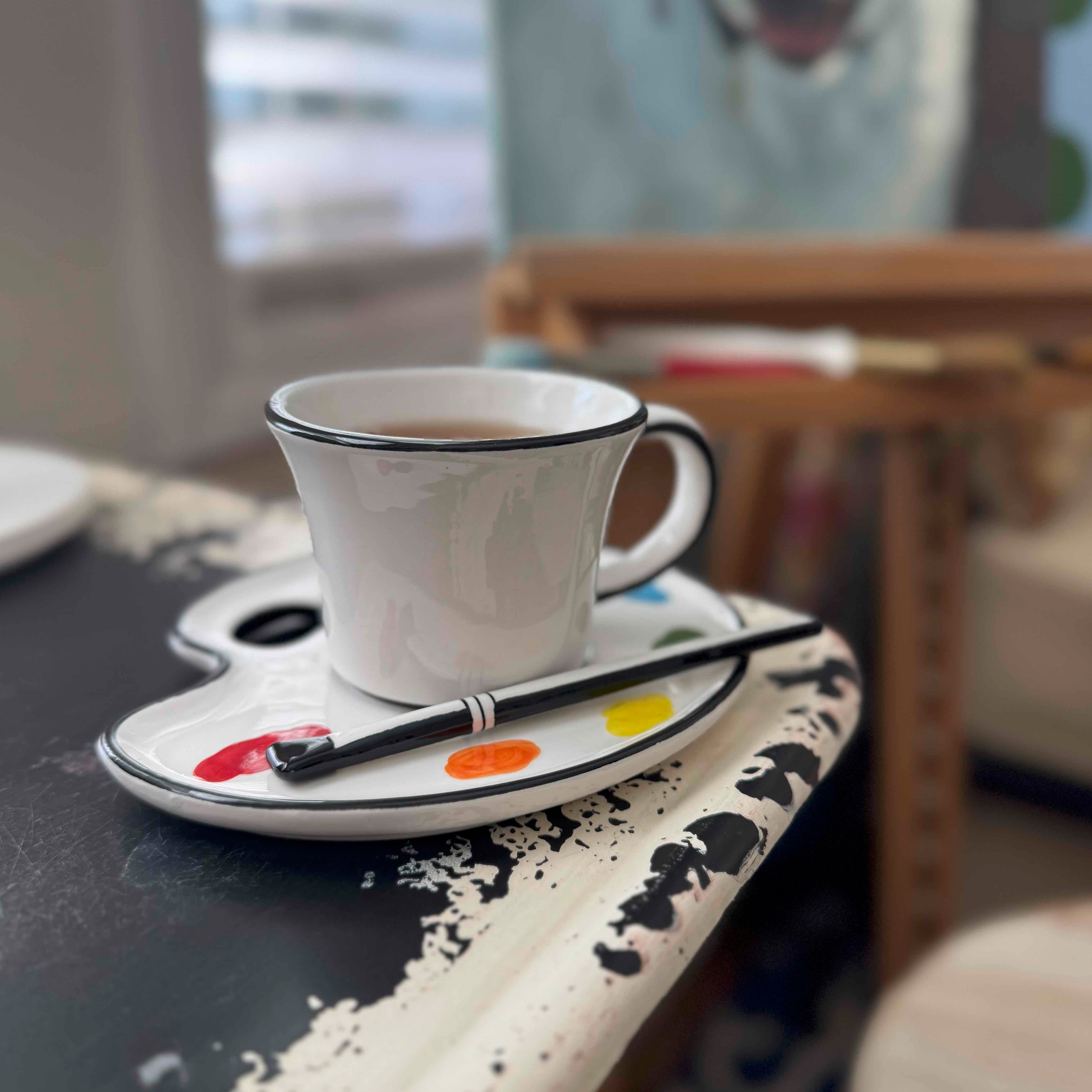 White mug with black rim on a saucer with paintbrushes, placed on a surface with artistic elements.