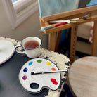 Artistic setup with a cup of tea, paintbrushes, and color palette on a table.