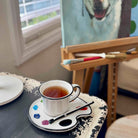 Cup of tea on a saucer with a paint palette and brush, next to an easel with a dog painting.