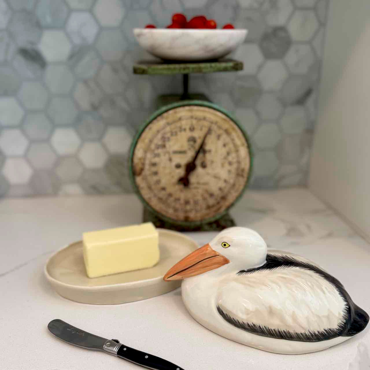 Butter dish shaped like a pelican with a bar of butter, knife, and vintage scale in the background.