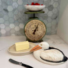 Butter dish shaped like a pelican with a bar of butter, knife, and vintage scale in the background.