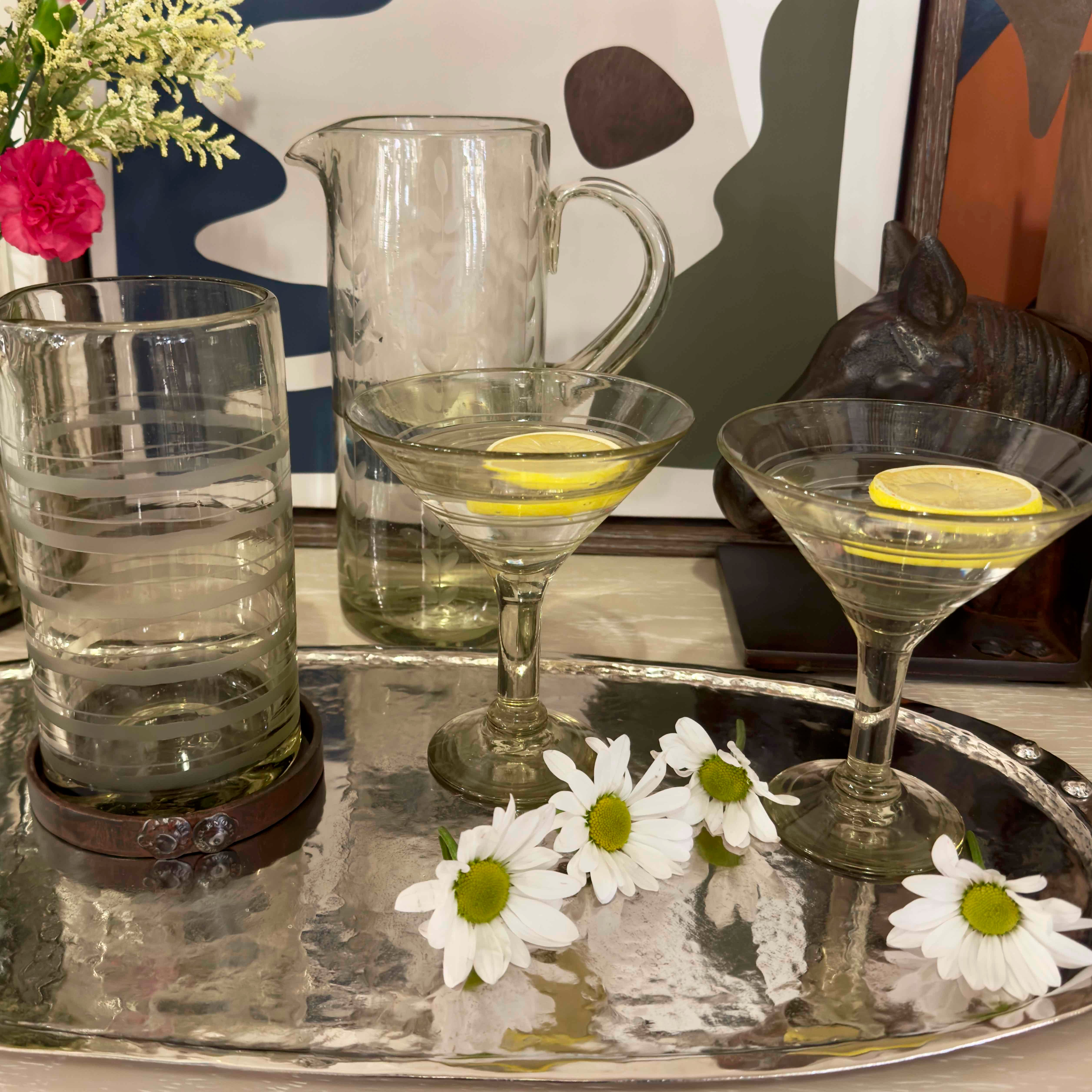 Two martini glasses with lemon slices on a silver tray with flowers and a pitcher in the background.