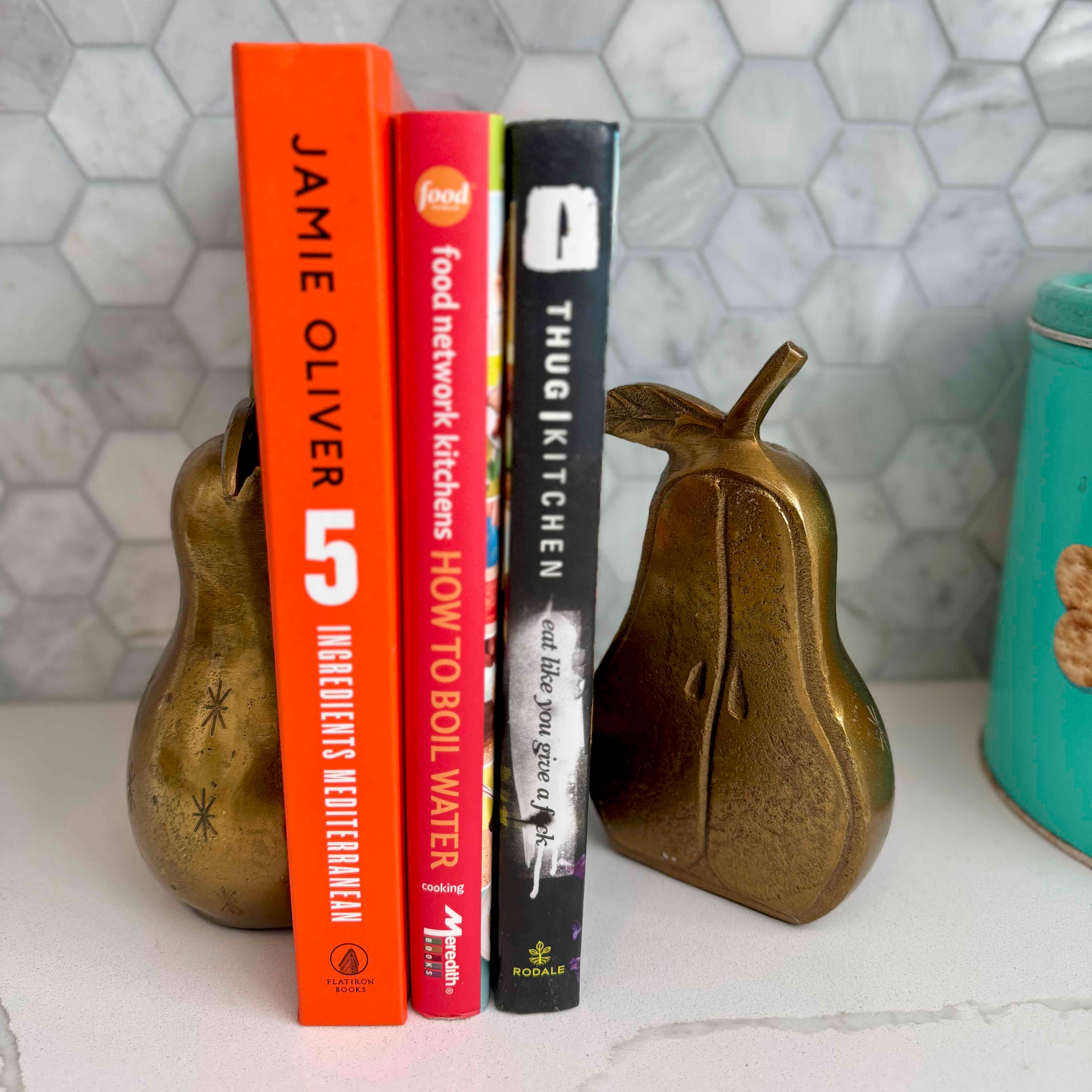 Three books on a surface with decorative pears and a teal container against a tiled wall.