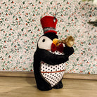 A plush toy figurine of a penguin wearing a red hat and a black coat with white polka dots, holding a trumpet, standing against a floral background.