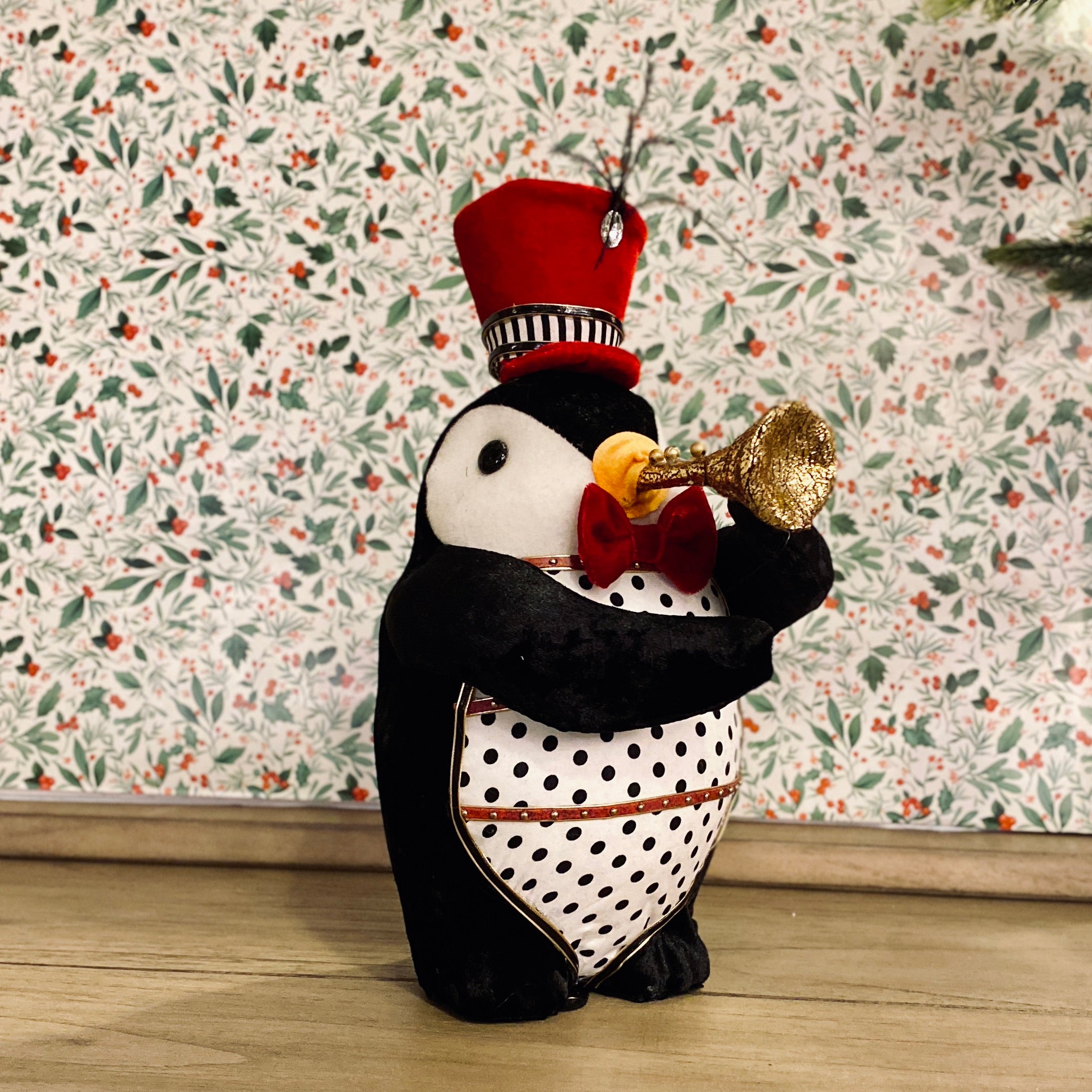 A plush toy figurine of a penguin wearing a red hat and a black coat with white polka dots, holding a trumpet, standing against a floral background.