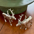 Two silver deer figurines on a wooden surface with a green container in the background.