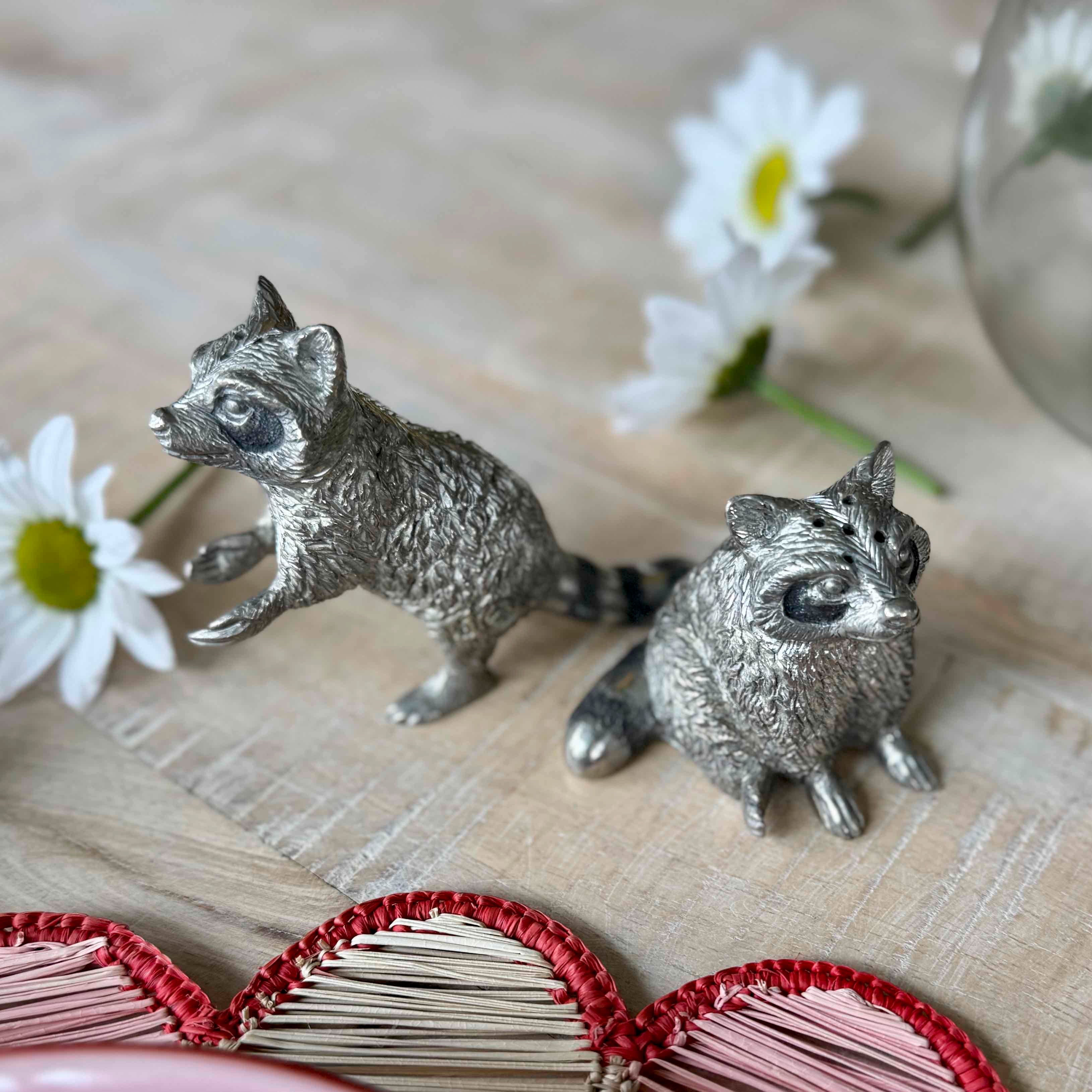 Two silver raccoon figurines on a textured surface with flowers in the background