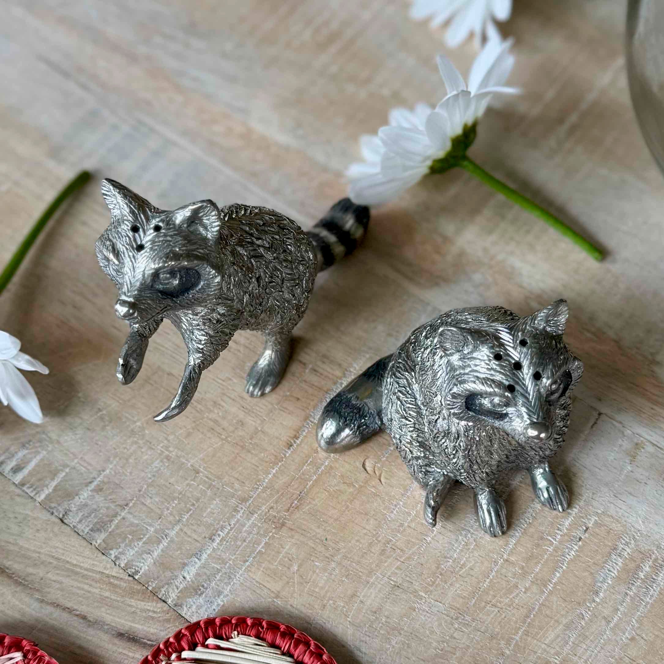 Two silver raccoon figurines on a textured surface with flowers in the background