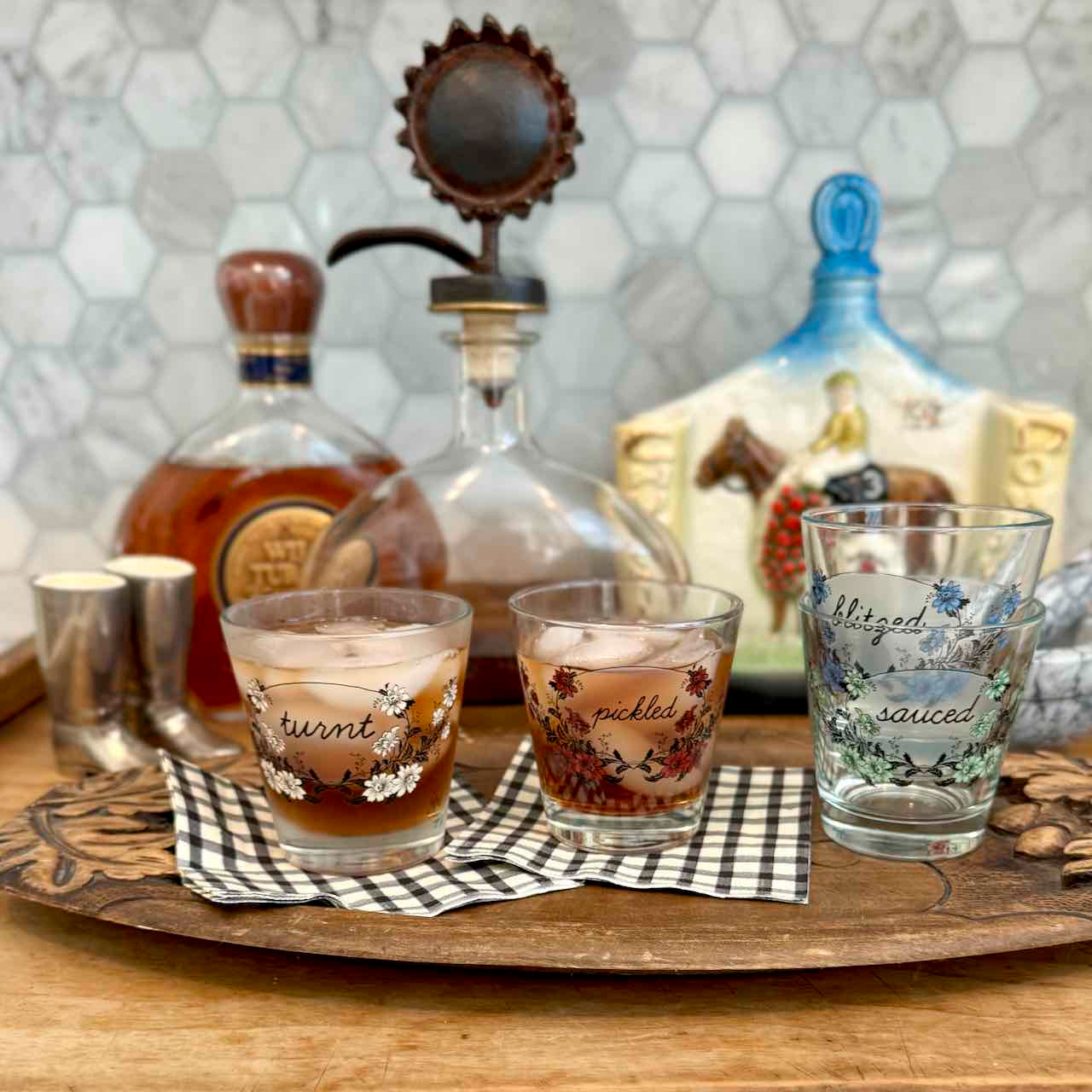 Three decorated clear drinking glasses with floral and script designs sit on a wooden tray in a kitchen setting, with bottles and a ceramic pitcher in the background.