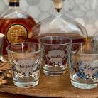 Three clear glass rocks tumblers with floral illustrations and handwritten labels are arranged on a wooden tray, with whiskey bottles in the blurred background.