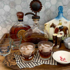 A styled bar scene with two frosted glasses filled with iced whiskey, a clear decanter, two small etched glasses, metal jiggers, a small white dish, and decorative bottles on a wooden tray against a tiled wall.