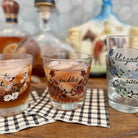 Three clear glass rocks tumblers with floral and script labels, shown filled with amber cocktails on a wooden surface with a checkered napkin and blurred bottles in the background.