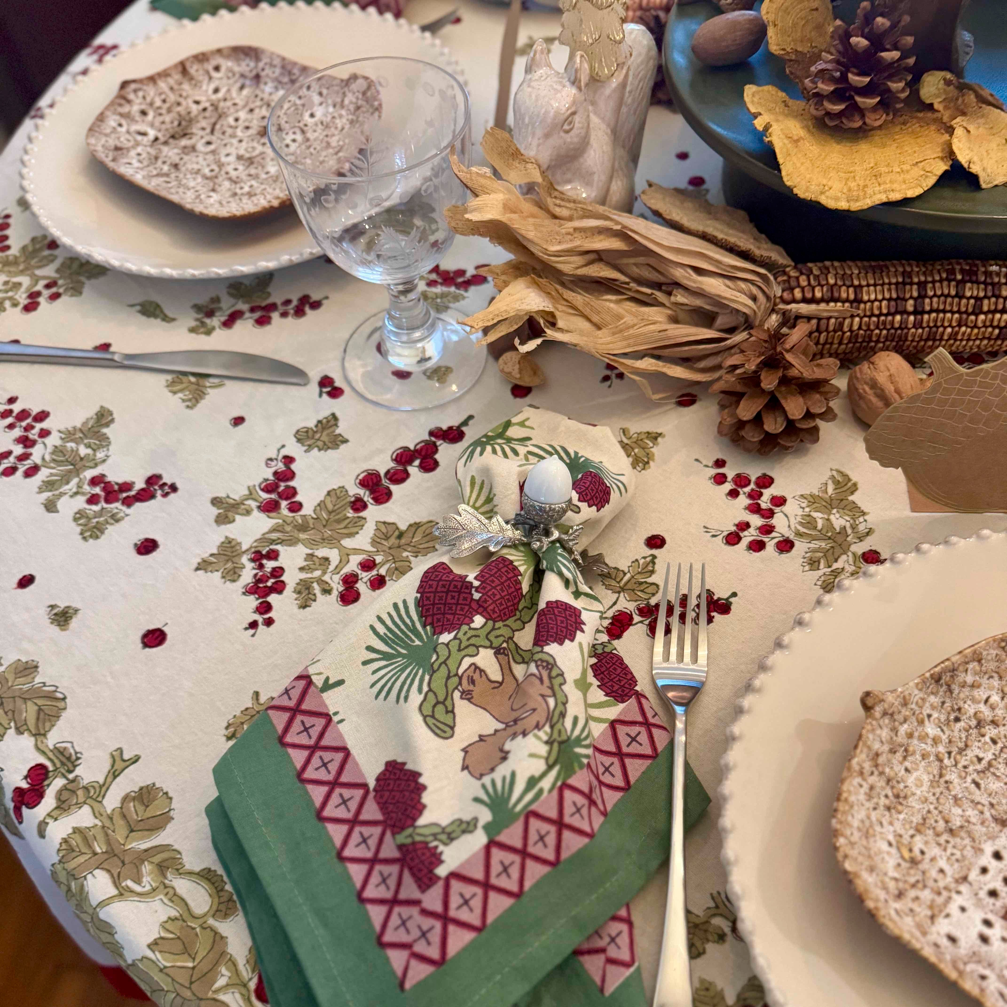 Set table with a decorative tablecloth featuring berries and leaves, glasses, plates, and cutlery.