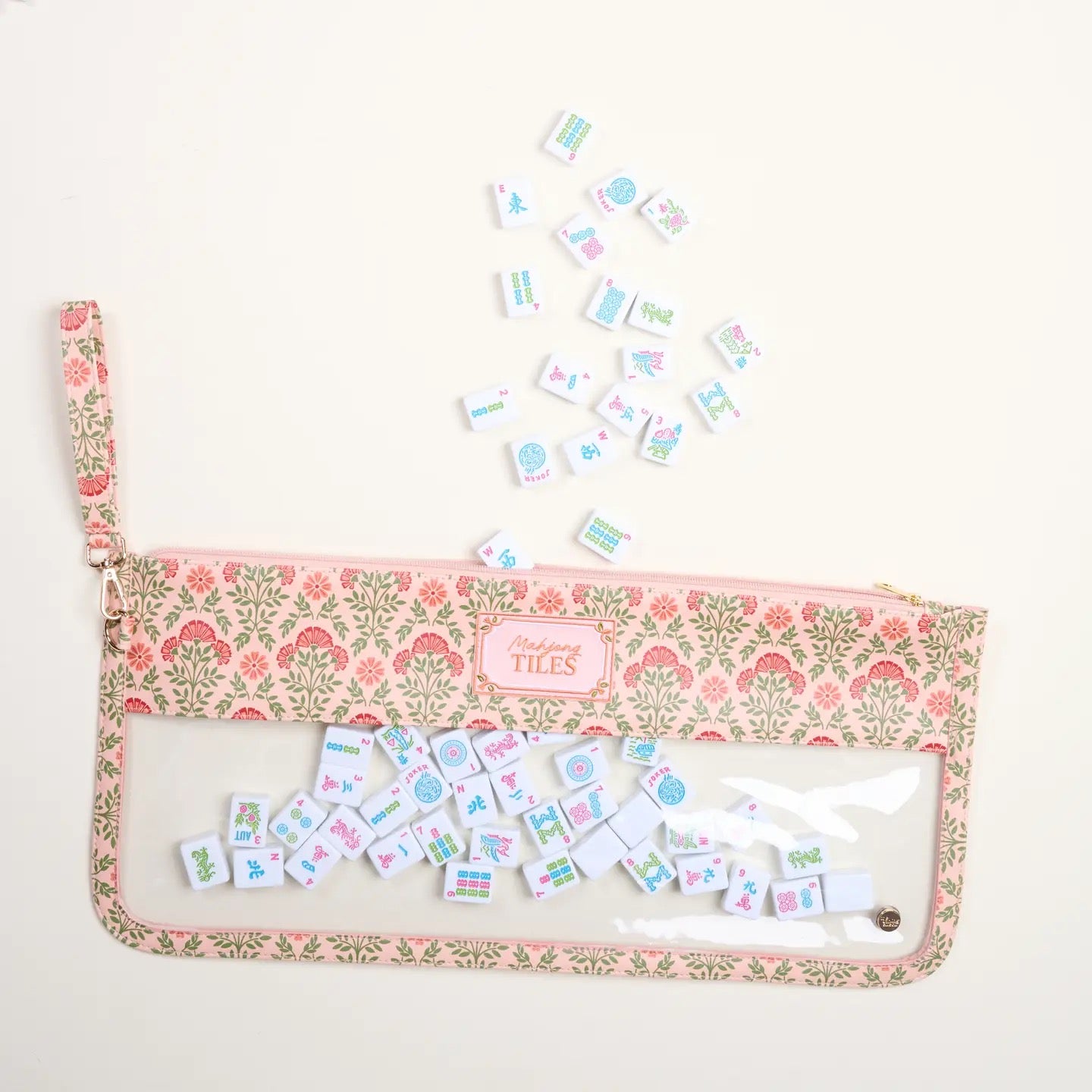 A clear zip pouch with a pink and green floral patterned fabric trim, featuring a small pink brand label that reads 'THE TIES'. The pouch is shown open with several small illustrated tiles scattered above and inside it, on a plain off-white background.