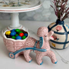 Pink ceramic poodle figurine with a cart full of colorful candies, placed on a surface with decorative items in the background.
