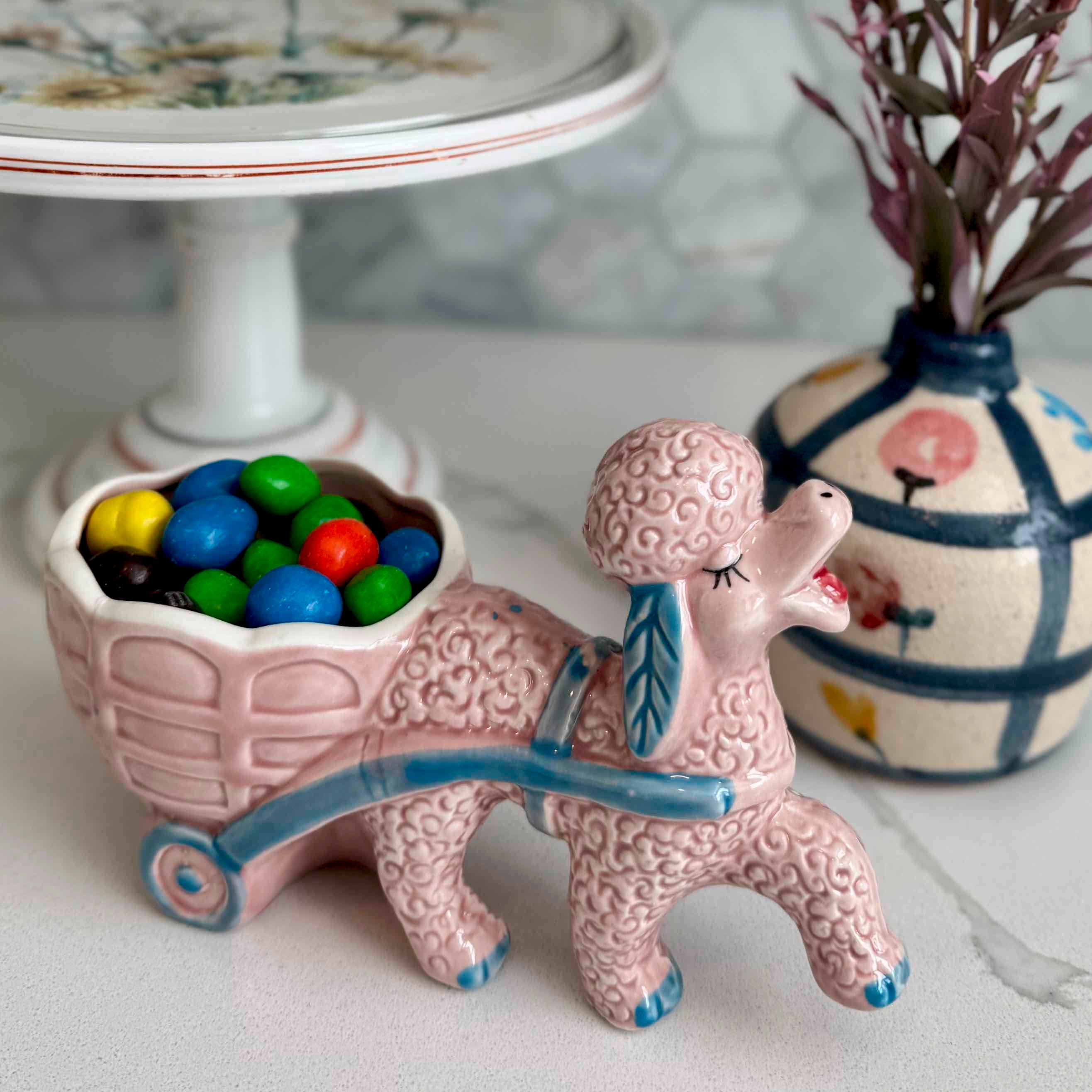 Pink ceramic poodle figurine with a cart full of colorful candies, placed on a surface with decorative items in the background.