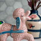 Pink ceramic poodle figurine with a brain on its head, standing in front of a decorative vase.