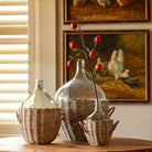 Three woven wicker baskets holding clear glass demijohn-style bottles, arranged on a wooden table in a warmly lit interior with framed chicken paintings on the wall behind.