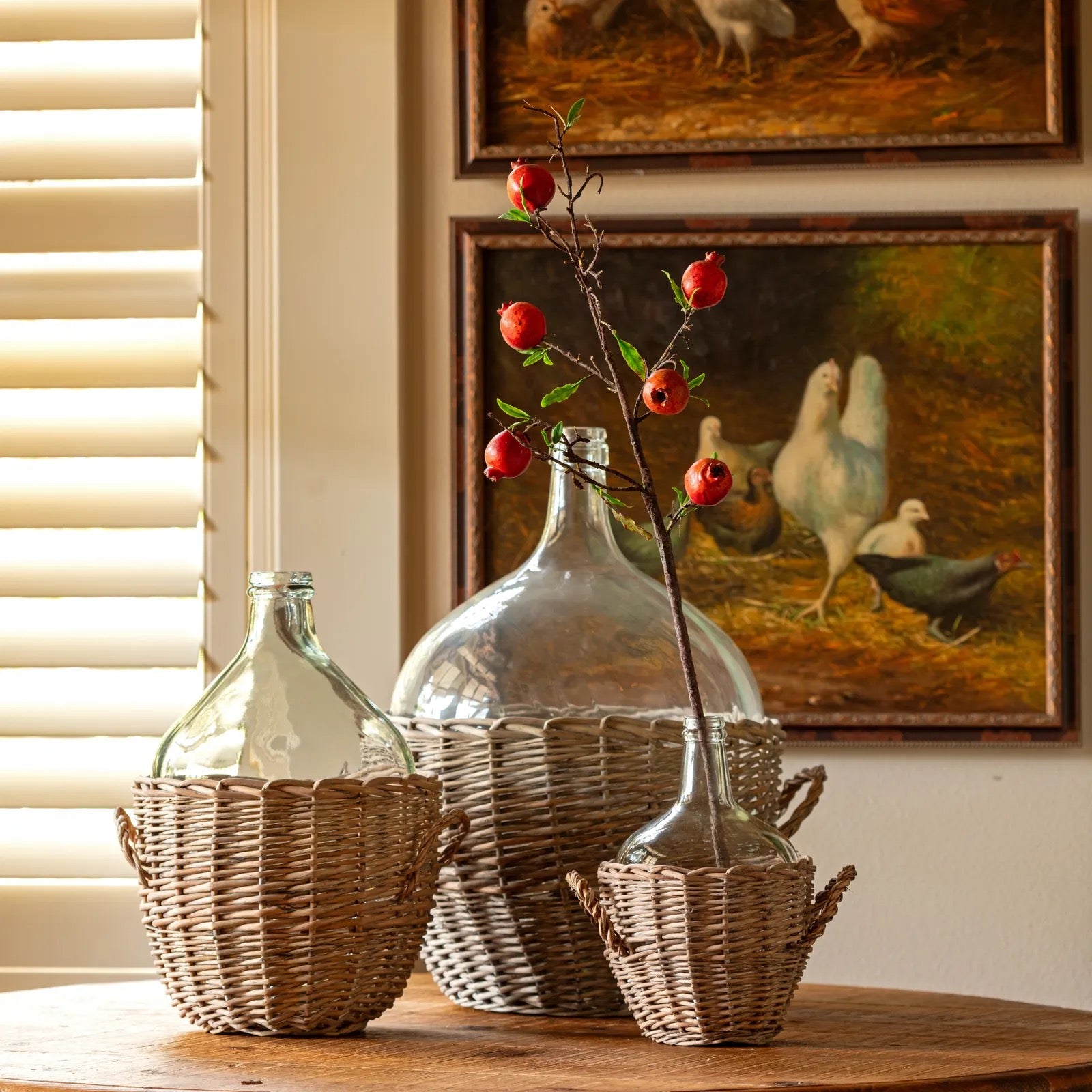 Three woven wicker baskets holding clear glass demijohn-style bottles, arranged on a wooden table in a warmly lit interior with framed chicken paintings on the wall behind.