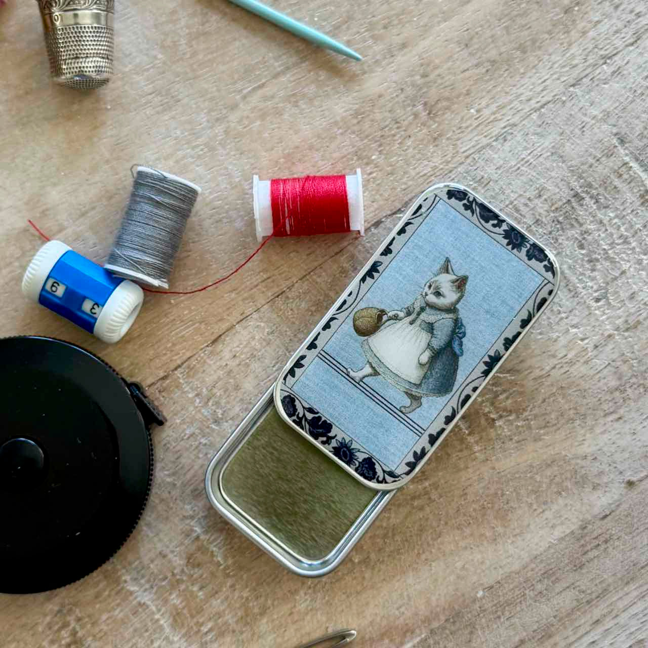A vintage-style metal tin with a printed illustration of a cat in a dress on the lid, placed on a wooden surface with sewing accessories nearby, including thread spools, a thimble, and a crochet hook.