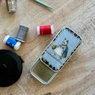 A vintage-style metal tin with a printed illustration of a cat in a dress on the lid, placed on a wooden surface with sewing accessories nearby, including thread spools, a thimble, and a crochet hook.