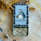 Tin box with decorative lid featuring a cat illustration on a wooden surface with sewing tools.