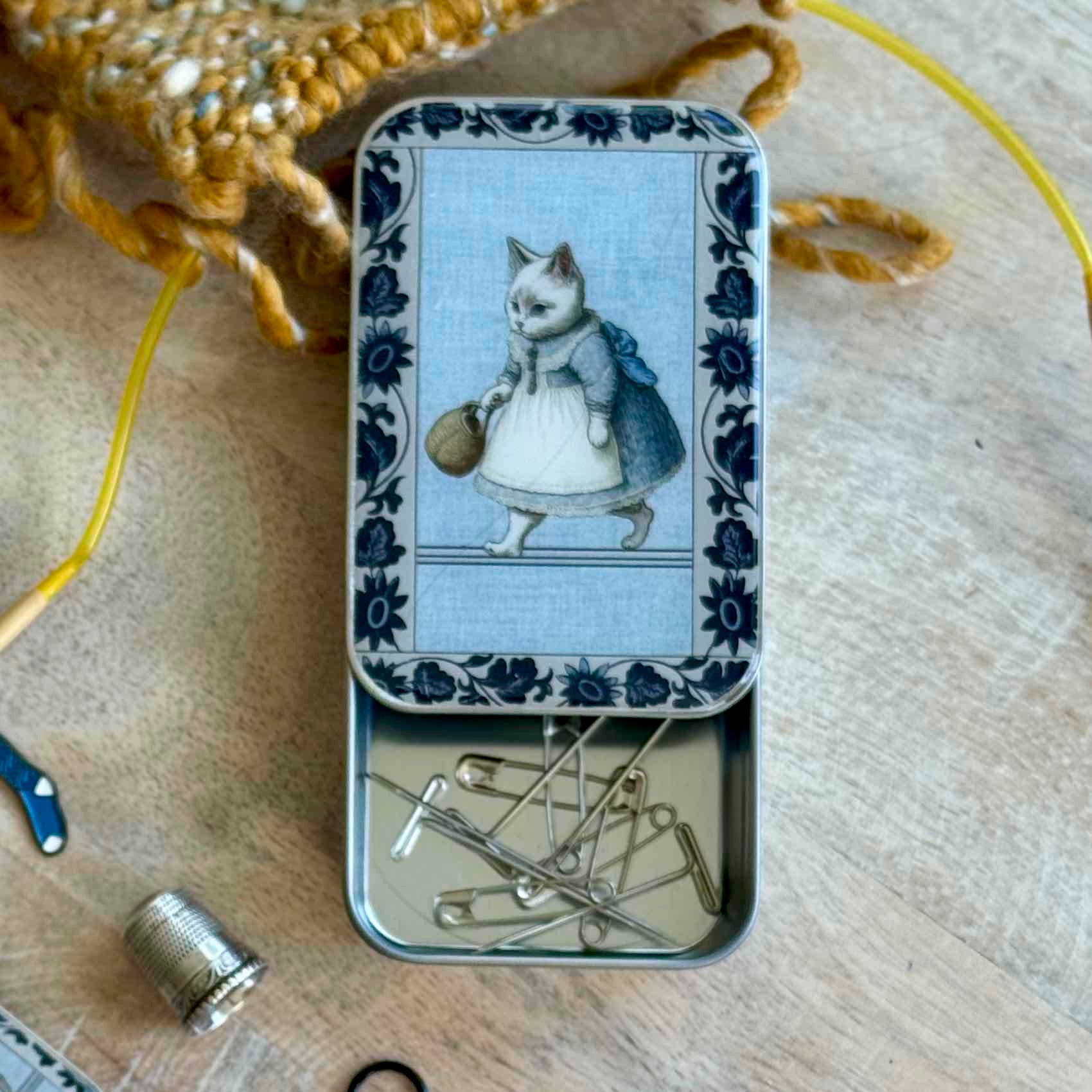 Tin box with decorative lid featuring a cat illustration on a wooden surface with sewing tools.