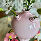 Pink ceramic vase with three small necks holding flowers, blurred floral background