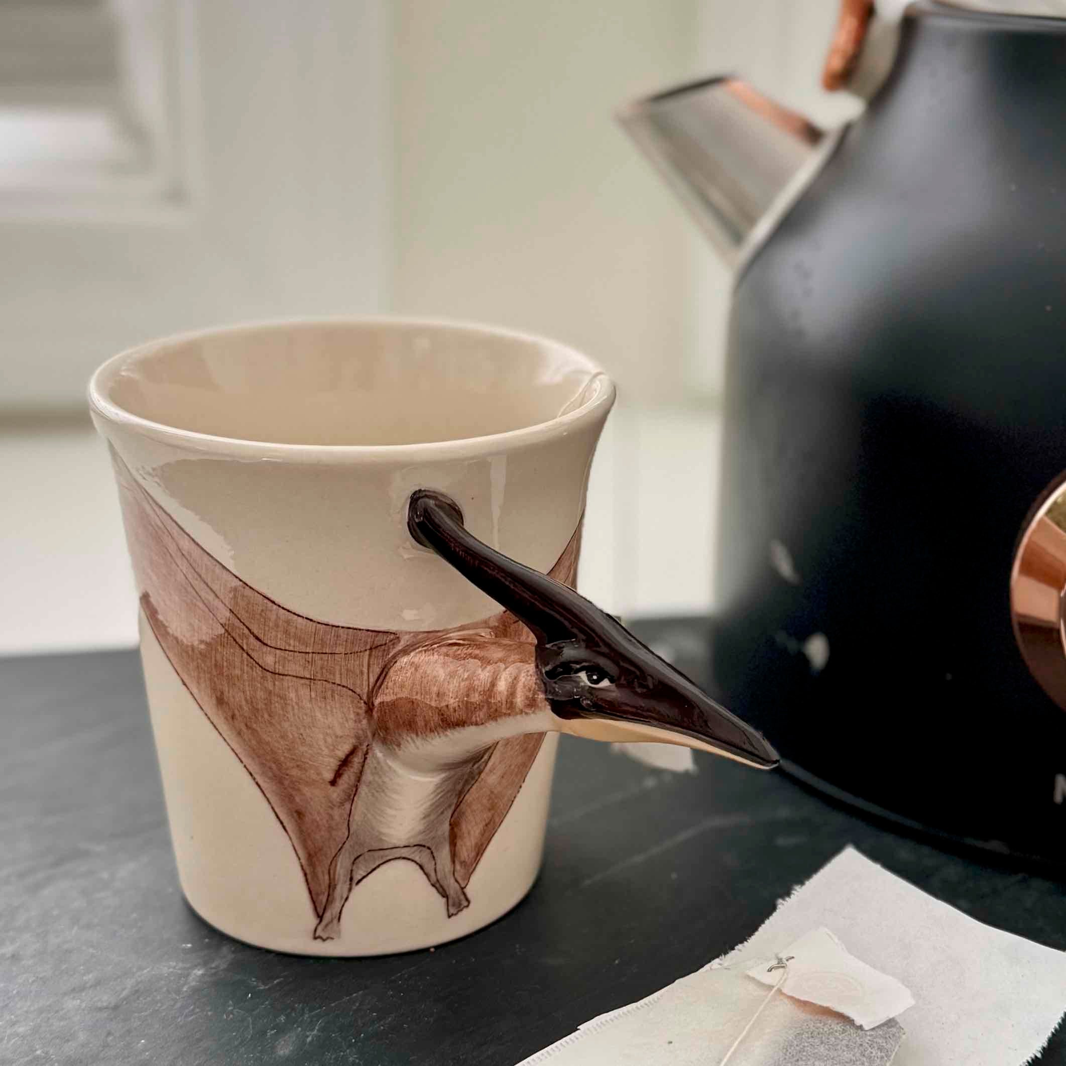 Ceramic cup with a bird design next to a black kettle on a surface.
