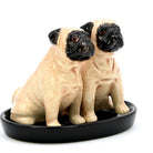 Two ceramic pug figurines sitting side by side on a black oval dish, photographed against a plain white background.
