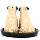 A pair of ceramic pug-shaped salt and pepper shakers sitting side-by-side on a round black dish, photographed against a solid white background.