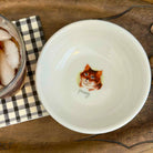 A white ceramic bowl with a hand-painted orange tabby cat illustration and the word 'pussy' written below it, shown on a wooden surface next to a checkered napkin and a glass of iced coffee.