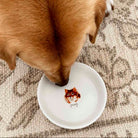 A golden-brown dog drinking from a small white ceramic bowl decorated with a custom illustrated cat face and the name 'puppy' underneath, placed on a patterned beige rug.