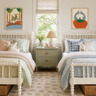 A styled children's bedroom featuring two matching white spindle-top beds with pastel bedding, a sage green nightstand, and framed abstract art on the walls.