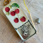 Small tin with embroidered design of raspberries and earrings on a wooden surface