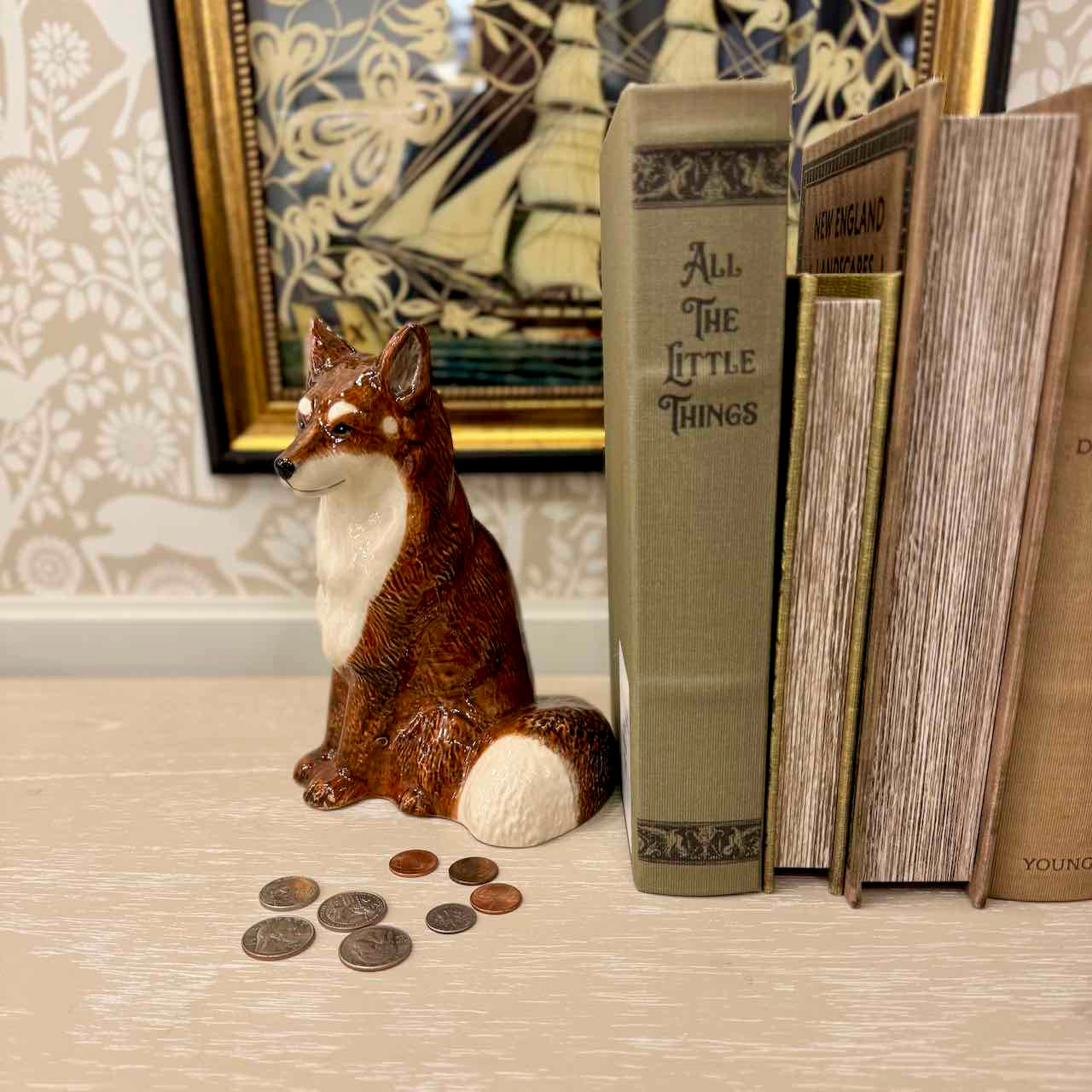 Fox figurine with coins next to books titled 'All The Little Things' and 'Young' on a decorative surface.