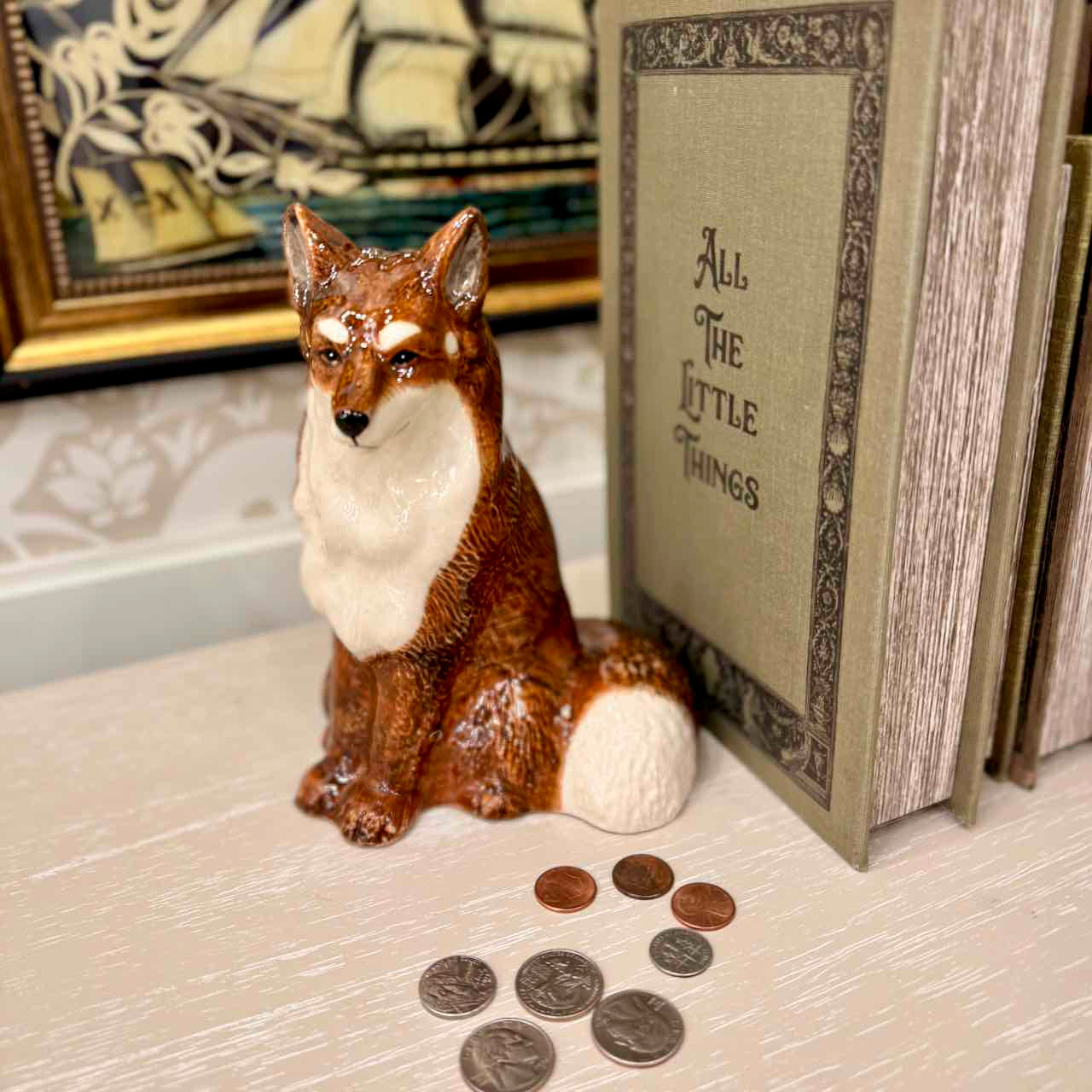 Fox figurine with coins on a surface next to a book titled 'All The Little Things'.