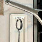 A white ceramic spoon rest with a black illustration of a spoon and the words 'Best in Grease' printed on it, placed on a light-colored countertop next to a stainless steel pan.