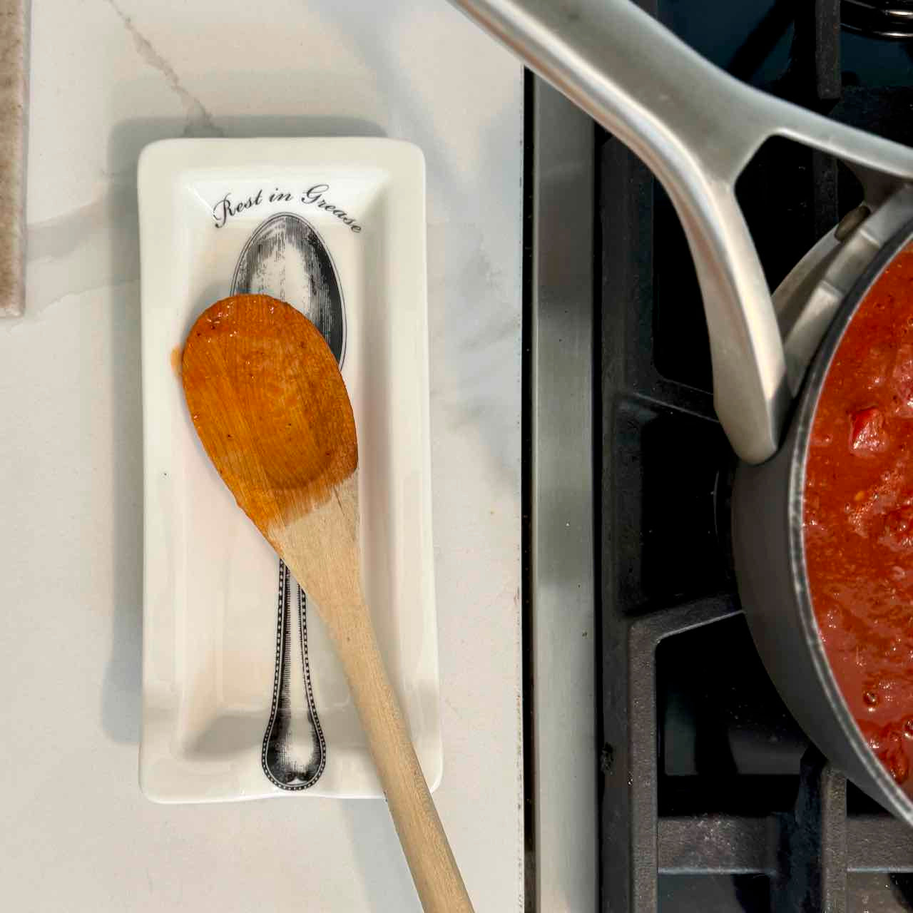 A wooden spoon rests on a white rectangular dish with a printed silver spoon and the words 'Real in Rome' on a light countertop beside a stove and a pot of red sauce.