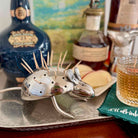 Metallic hedgehog-shaped drink stirrer on a bar tray with bottles and a glass in the background.