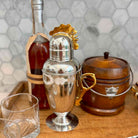 A polished silver cocktail shaker with a gold-toned rooster handle and lid ornament, displayed on a wooden surface alongside a glass, a brown bottle, and a wooden ice bucket against a tiled wall.