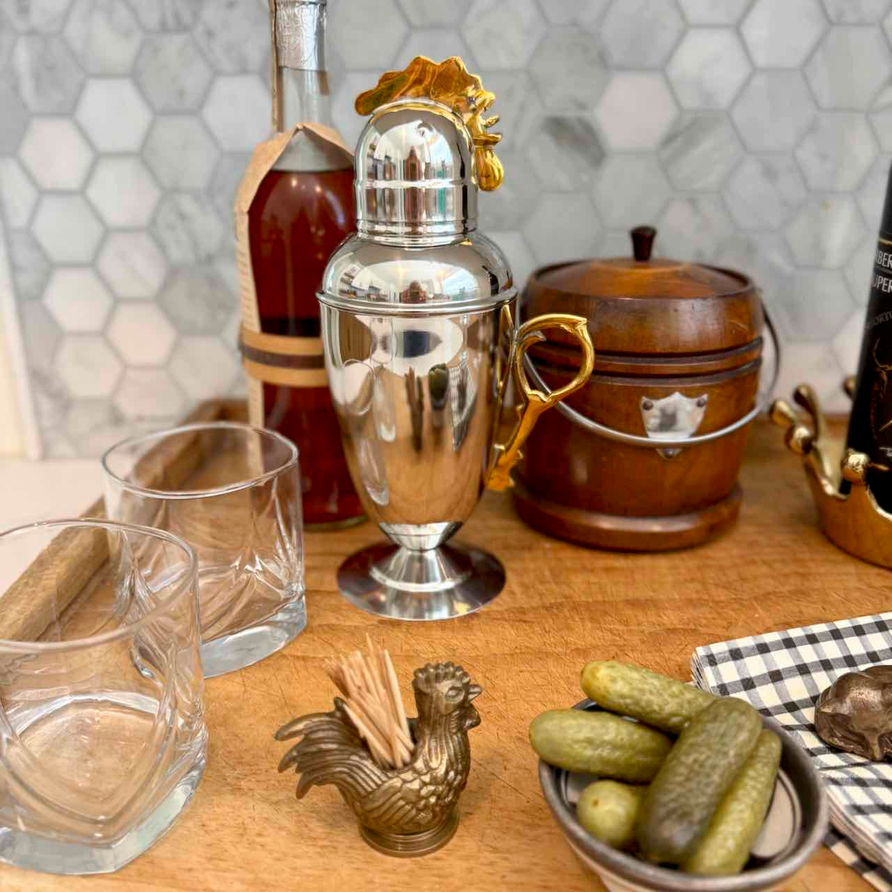 A polished silver cocktail shaker with a gold rooster topper sits on a wooden bar surface, surrounded by bar accessories including two clear lowball glasses, a small brass toothpick holder shaped like a rooster, pickles in a bowl, and a dark bottle in the background.