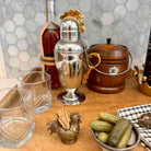 A polished silver cocktail shaker with a gold rooster topper sits on a wooden bar surface, surrounded by bar accessories including two clear lowball glasses, a small brass toothpick holder shaped like a rooster, pickles in a bowl, and a dark bottle in the background.