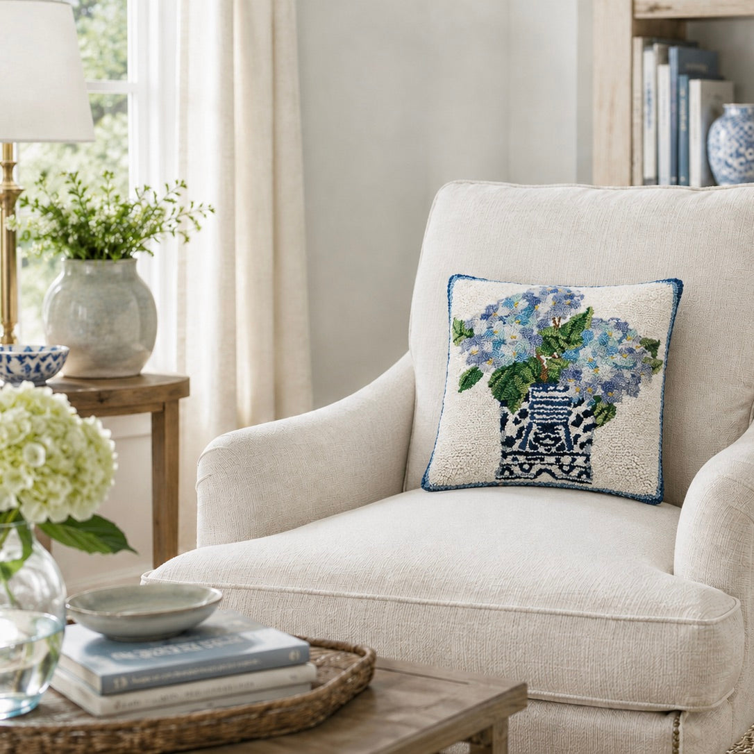 A square needlepoint throw pillow featuring blue and white hydrangeas in a blue patterned vase, styled on a cream-colored armchair in a bright, neutral-toned living room.