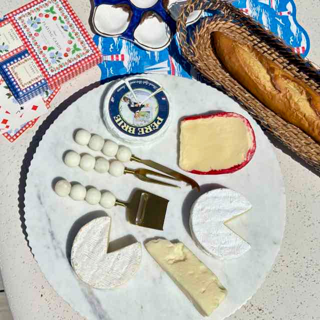 A styled cheese and bread charcuterie board arranged on a round white marble platter, with additional patterned napkins and a woven bread basket in the background.