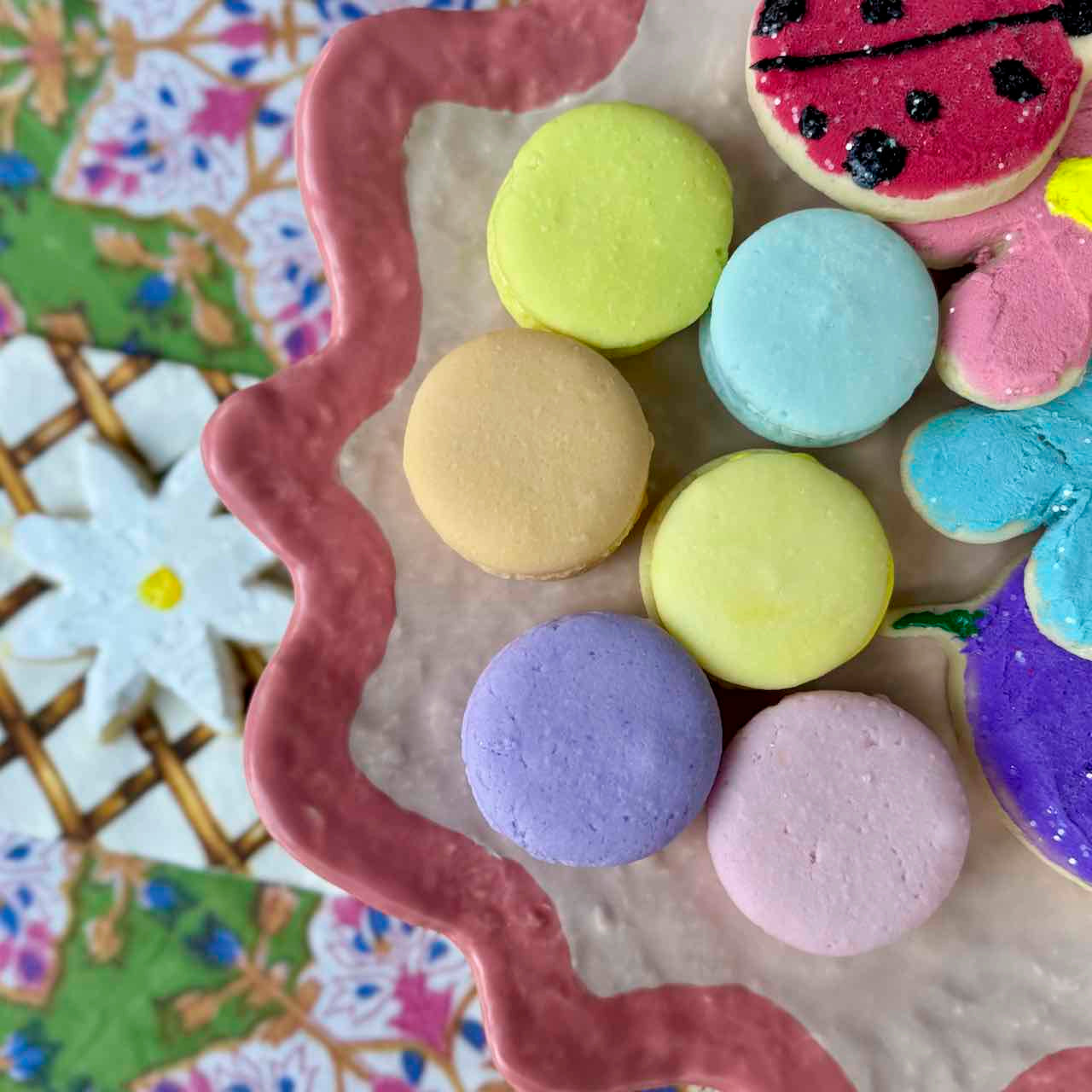 A close-up of pastel macarons and decorated cookies arranged on a pink scalloped plate, with a floral-patterned surface visible in the background.
