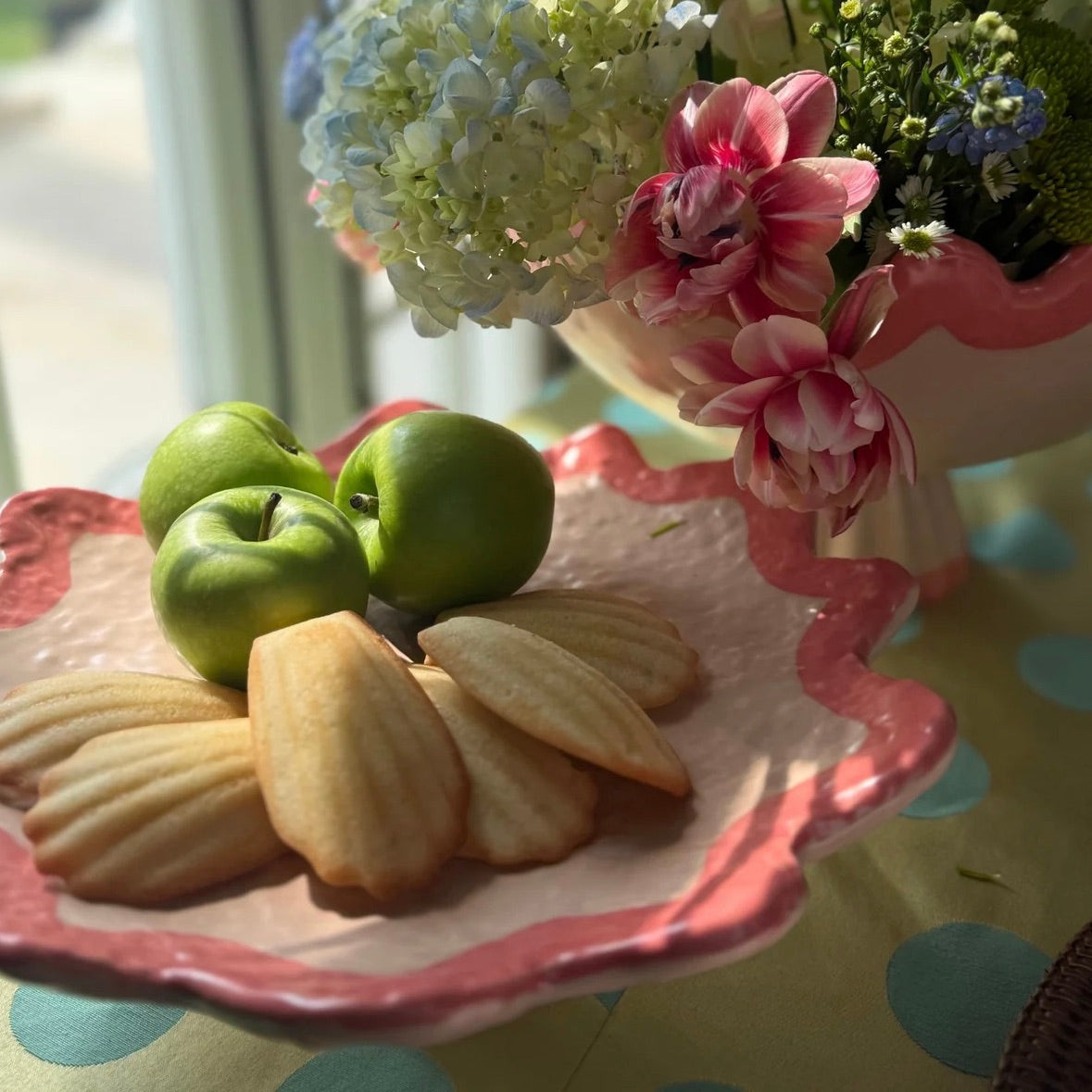 A pink and white scalloped ceramic serving platter on a table, holding three green apples and several pale baked pastries, with flowers in the background near a window.