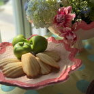 A pink and white scalloped ceramic serving platter on a table, holding three green apples and several pale baked pastries, with flowers in the background near a window.