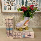 Decorative setup with books, a ceramic shoe with wheels, and flowers against a patterned wall.